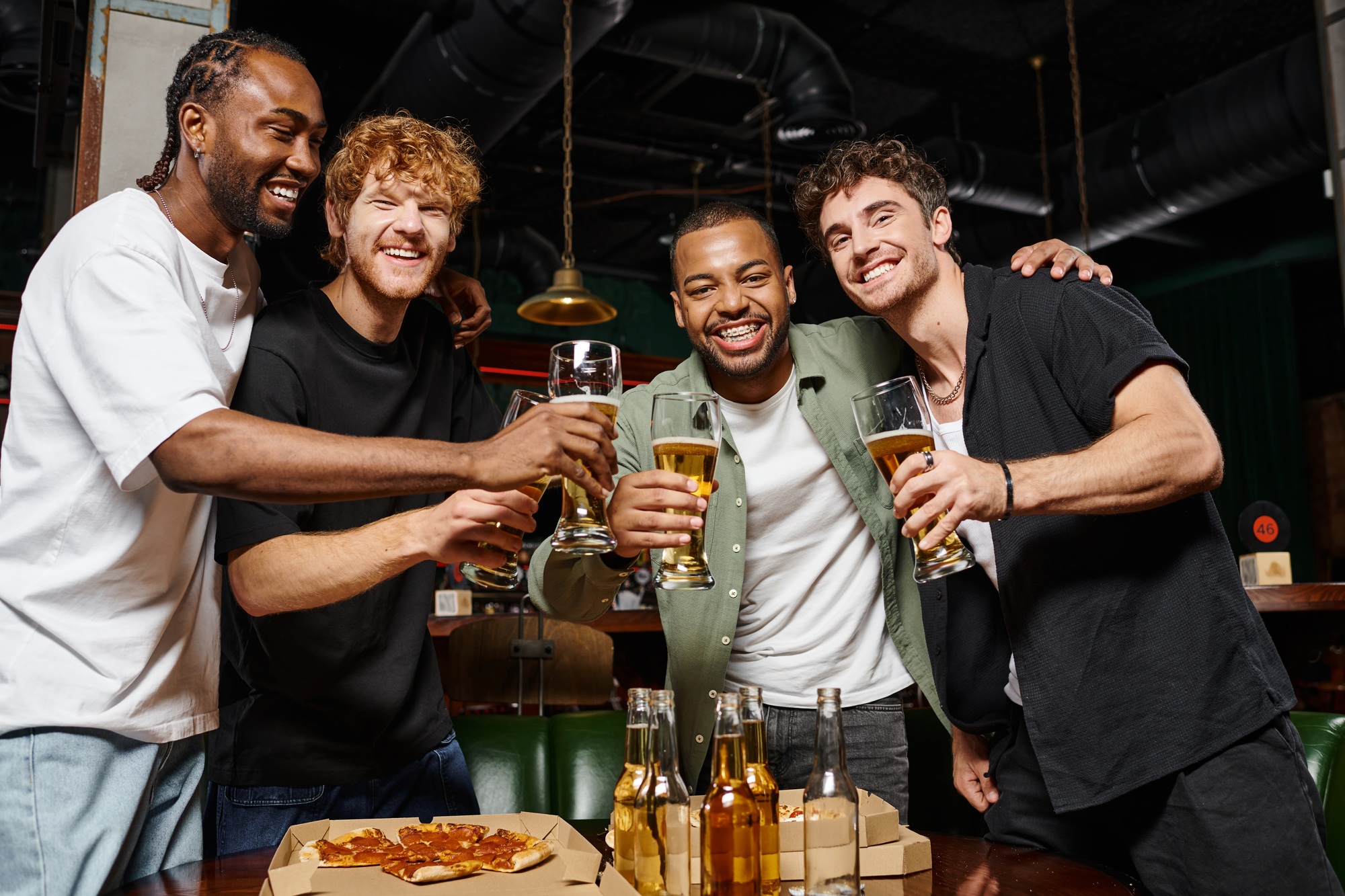 happy interracial men toasting with glasses of beer in bar during bachelor party, male friendship
